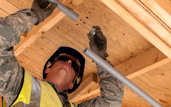 A military service member wearing US Air Force digital camouflage (ABU pattern) and a high-visibility safety vest is installing conduit piping in the ceiling joists of a wood-framed structure under construction. The individual is equipped with personal protective equipment including a hard hat, safety glasses, and work gloves. This appears to be a military engineering or civil engineering construction project.