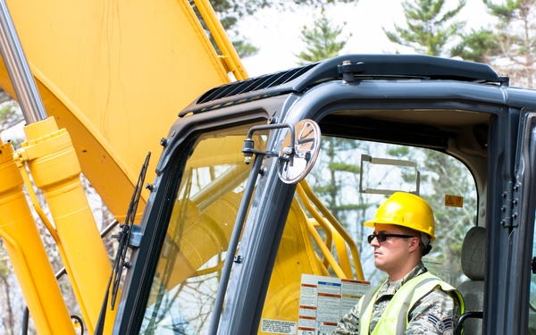 A military service member in camouflage uniform operates a yellow excavator or heavy construction equipment. The operator is wearing a yellow hard hat, safety glasses, and a high-visibility green vest over their military fatigues. The setting appears to be a wooded area, suggesting a military engineering or civil construction operation.