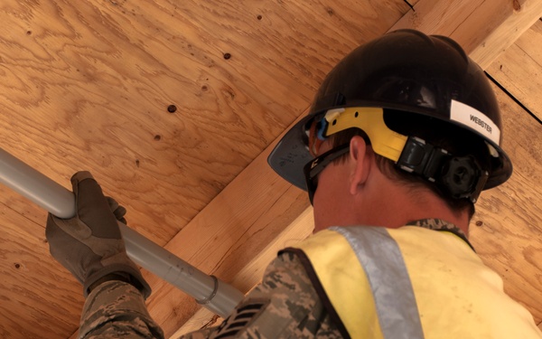 A military service member wearing camouflage uniform, black hard hat labeled 'WEBSTER', and a high-visibility yellow safety vest is installing or routing conduit along a plywood ceiling in a construction setting. The individual is working overhead, handling a gray metal or PVC conduit pipe while equipped with work gloves and a tool belt. The image is taken from a low angle looking upward, providing a close-up perspective of the work being performed.