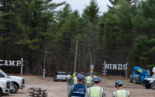 Military personnel wearing camouflage uniforms and high-visibility safety vests are walking through the entrance of Camp Hinds, a Boy Scouts of America camp. The scene suggests a work detail or disaster relief/cleanup operation, with construction equipment and multiple white utility trucks visible on the grounds. The camp entrance is marked by large rustic wooden letters spelling 'CAMP HINDS' flanked by pine forest.