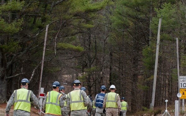 A group of military personnel wearing high-visibility yellow safety vests and blue hard hats are walking down a dirt road entering Camp Hinds, a Boy Scouts of America facility operated by the Pine Tree Council. The entrance is marked by large rustic wooden signs reading 'CAMP' and 'HINDS' on either side of the road. The setting appears to be early spring based on the sparse foliage on the deciduous trees.