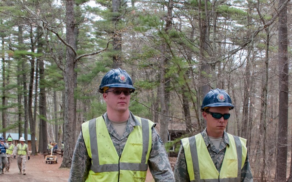 A group of military personnel wearing camouflage uniforms, high-visibility safety vests, blue hard hats, and safety glasses are walking along a dirt road through a forested area. The two individuals in the foreground appear to be leading a larger group visible in the background. They appear to be conducting a cleanup or work detail operation, with at least one person carrying a black trash bag.