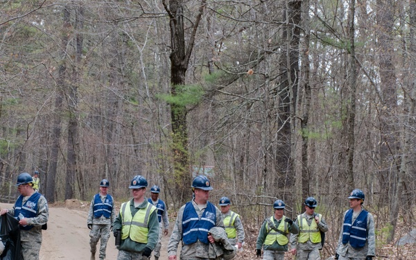 A group of military personnel in camouflage uniforms are walking along a dirt road through a wooded area. They are wearing blue hard hats and high-visibility safety vests, suggesting an organized work or cleanup detail. A white pickup truck is parked on the left side of the road.