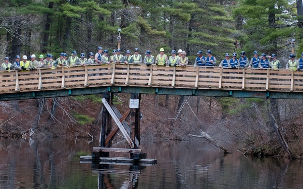 A large group of construction or safety workers wearing hard hats and high-visibility vests are gathered on a newly constructed wooden pedestrian bridge over a calm body of water. The bridge appears to be a recent build or renovation, featuring fresh lumber railings atop an older steel/timber substructure. The still water below creates a clear mirror-like reflection of the bridge and the workers standing on it.