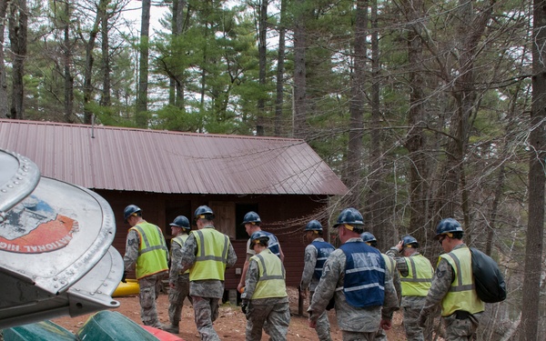 A group of approximately 12 personnel in military camouflage uniforms, wearing blue hard hats and high-visibility yellow safety vests, are walking in a line toward a small red-roofed wooden cabin in a forested area. Stacked canoes in silver, green, and red are visible in the foreground left. The setting appears to be a camp or outdoor training facility near a body of water.