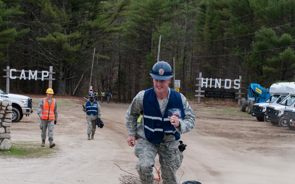 A person in military camouflage uniform wearing a blue hard hat and blue safety vest is running or jogging out of the entrance to 'Camp Hinds,' a Boy Scouts of America camp. They appear to be dragging something behind them on a rope. Several other personnel in uniform and safety gear are visible in the background near vehicles and equipment.