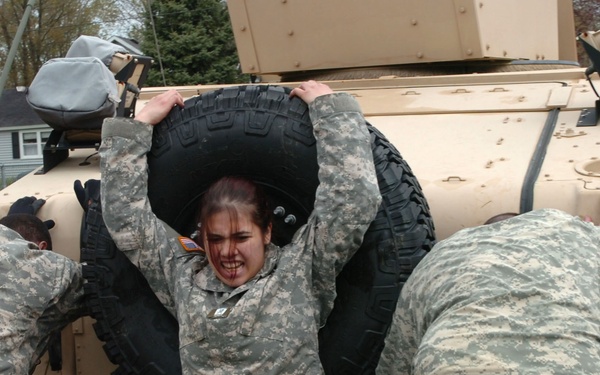Three soldiers in US Army ACU (Army Combat Uniform) digital camouflage are engaged in a physical training or evaluation exercise involving a military vehicle, likely a HMMWV (Humvee). The central soldier, a female, is visibly straining as she lifts or holds a large spare tire overhead with an intense expression. The other two soldiers are pushing against the sides of the vehicle.