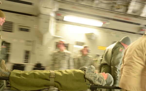 Military personnel are carrying a person on a stretcher through what appears to be the interior of a large military transport aircraft. The scene depicts an aeromedical evacuation or medical training exercise, with multiple uniformed individuals moving urgently through the cargo bay. Motion blur in the image suggests rapid movement and urgency.