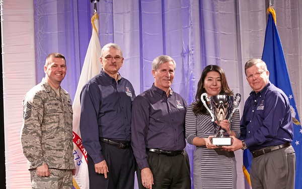 Five individuals are posed on a stage during what appears to be a formal awards ceremony. One person in military camouflage uniform stands alongside four civilians, one of whom is holding a silver trophy cup. The setting includes American and Air Force flags, suggesting a military-affiliated recognition event.