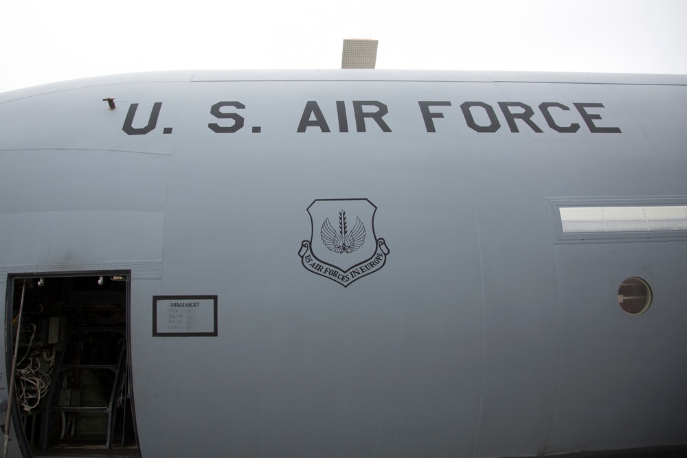 The image shows the fuselage of a U.S. Air Force military aircraft, prominently displaying the 'U.S. AIR FORCE' text and a shield emblem reading 'Air Forces in Europe'. An open access panel or door on the left side reveals internal wiring and components, with a labeled 'ARMAMENT' placard visible. The aircraft is painted in standard military gray livery.
