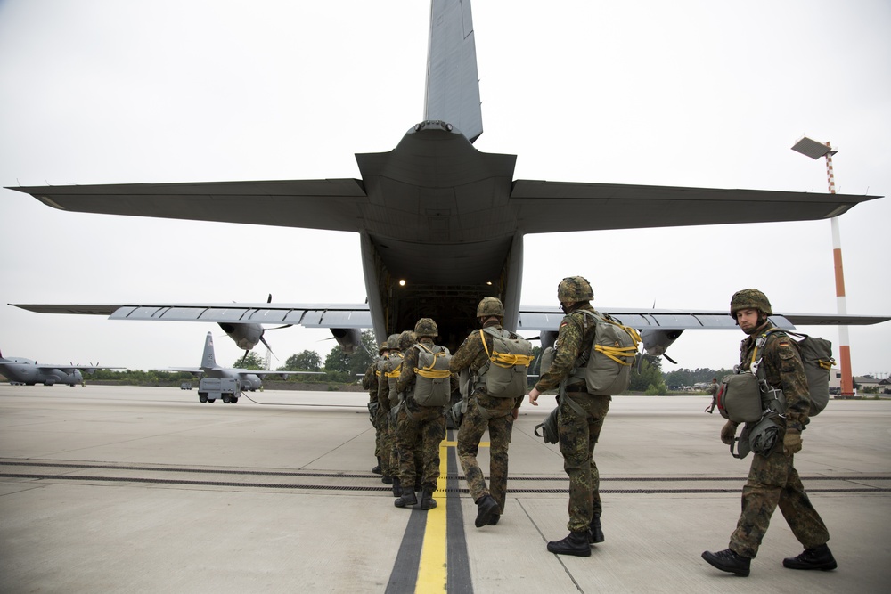 A group of military paratroopers in full combat gear are boarding a large military transport aircraft via its rear ramp on an airfield tarmac. The soldiers are equipped with parachute packs secured with yellow straps, helmets, and tactical backpacks. A second military transport aircraft is visible in the background on the left side of the frame.