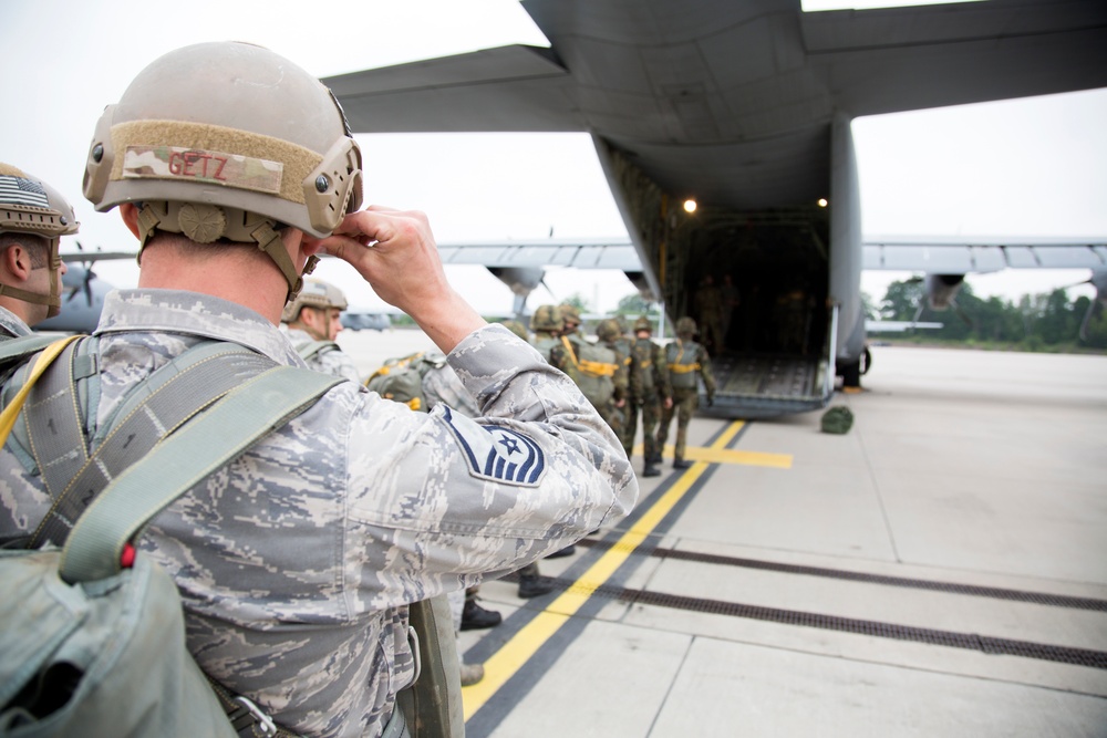 Military personnel in ABU (Airman Battle Uniform) camouflage are boarding or preparing to board a large military transport aircraft, likely a C-130 Hercules, via its rear ramp. The foreground soldier, identified by the name tape 'GETZ' on his helmet, wears Senior Master Sergeant rank insignia and is adjusting his combat helmet. The scene appears to be a pre-jump or airlift operation on a military airfield.
