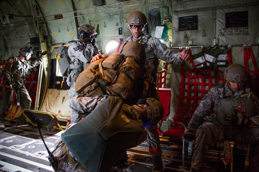 Military personnel are inside a military transport aircraft, appearing to be preparing for a parachute jump operation. Soldiers in full combat gear with helmets, parachute packs, and tactical equipment are visible in various stages of preparation. The interior of the aircraft shows characteristic red cargo netting, yellow safety markings, and standard military aircraft fittings.