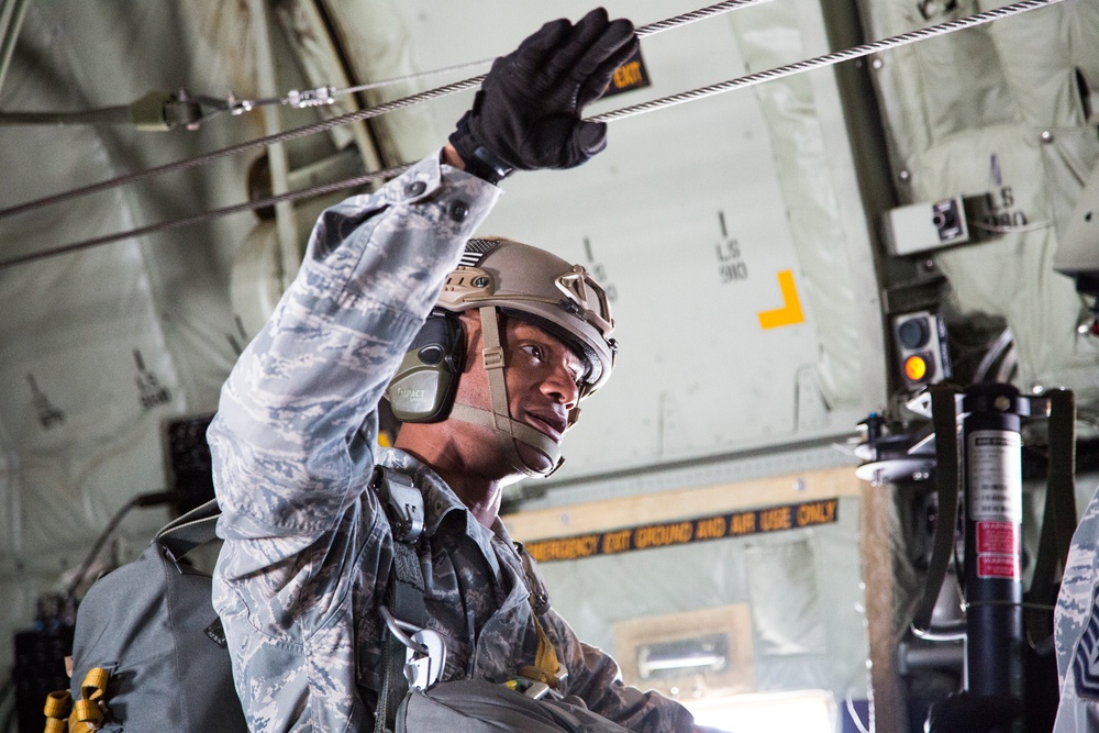 A U.S. Air Force service member in ABU (Airman Battle Uniform) is inside a military transport aircraft, reaching up to handle a static line cable. The individual is wearing a tactical helmet with hearing protection, tactical gloves, and appears to be equipped with a parachute harness. The setting is the interior of what appears to be a C-130 or similar military cargo/transport aircraft prepared for airborne operations.