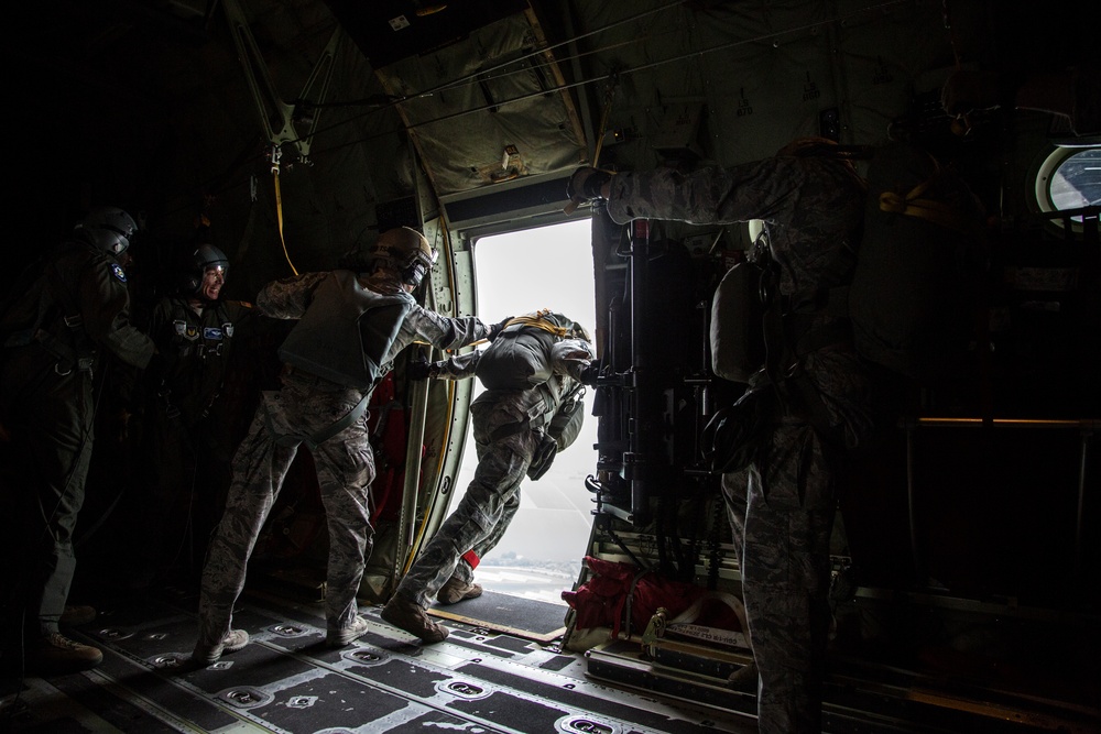 Military personnel are conducting a parachute jump operation from the open side door of a military transport aircraft, likely a C-130 or similar aircraft. One jumper is positioned at the door threshold, being assisted or checked by another service member, while additional personnel observe from inside the darkened cargo bay. The personnel are wearing military camouflage uniforms, helmets, and parachute equipment.