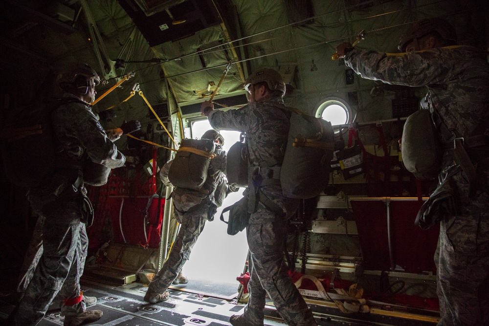 Military personnel in camouflage uniforms are preparing to conduct a parachute jump from the open rear ramp of a military transport aircraft, likely a C-130 Hercules. Jumpmasters are visible managing static lines attached to an overhead cable while jumpers are positioned near the open door. The scene is backlit by bright exterior light through the aircraft's open ramp.