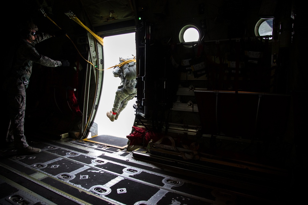 A military paratrooper is captured mid-jump exiting the side door of a military transport aircraft, likely a C-130 Hercules. A jumpmaster in camouflage uniform stands at the door observing the jump, with a static line visible connecting the jumper to the aircraft's anchor line cable. The interior of the aircraft is dark, contrasting sharply with the bright exterior sky visible through the open door.