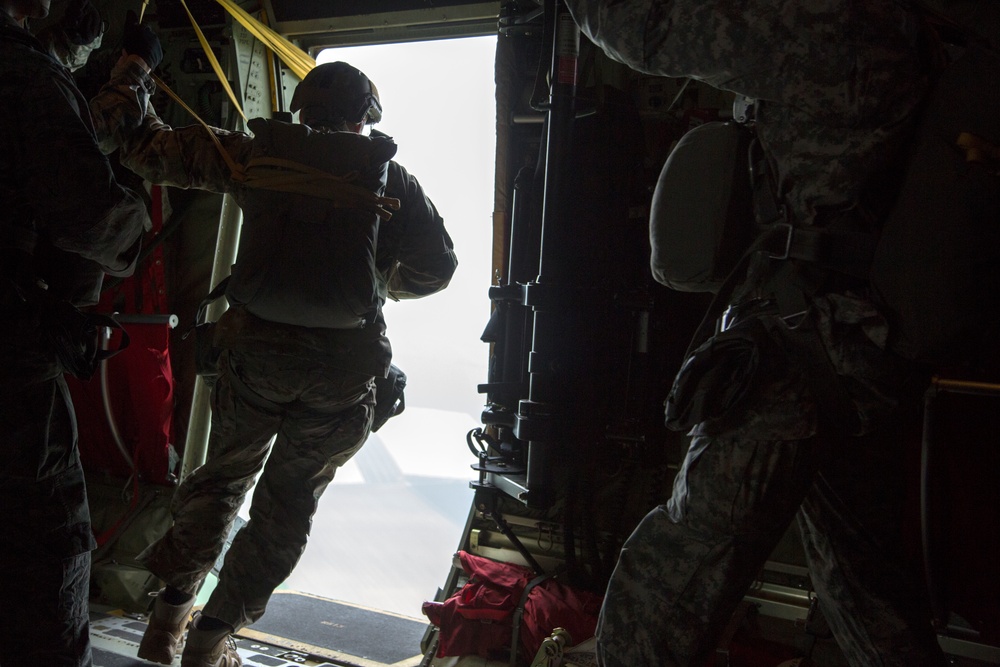Military personnel are positioned at the open rear ramp of a military transport aircraft, appearing to be preparing for or in the process of a parachute jump operation. The image is taken from inside the aircraft looking toward the open ramp door, with bright exterior light creating strong silhouettes of the soldiers. Red equipment bags are visible on the aircraft floor near the ramp.
