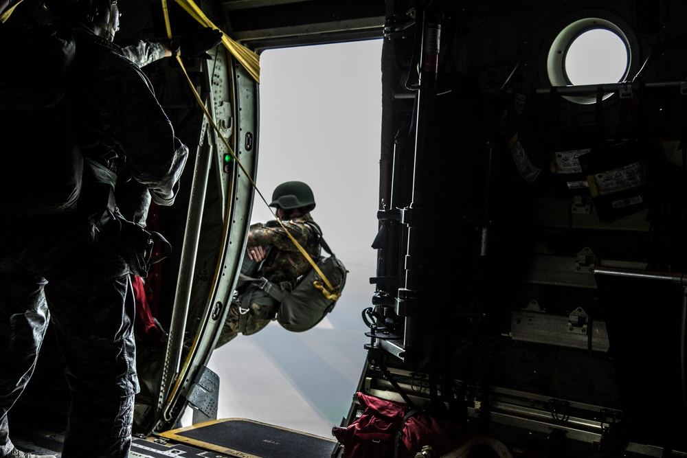 A military paratrooper is captured mid-exit from the open door of a military aircraft, likely a C-130 or similar transport plane. A static line is visibly attached, indicating a static-line parachute jump. Another service member stands inside the aircraft observing the jump.