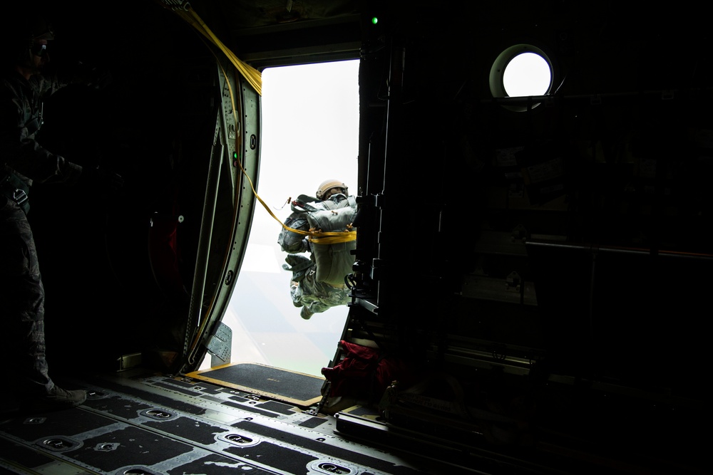 A military paratrooper is captured mid-exit from the open side door of a military transport aircraft, with a static line (yellow/orange cord) visibly attached and deployed. The image is taken from inside the aircraft's dark interior looking outward toward a bright, overcast sky. A second personnel member is visible in silhouette on the left side of the frame, likely a jumpmaster.