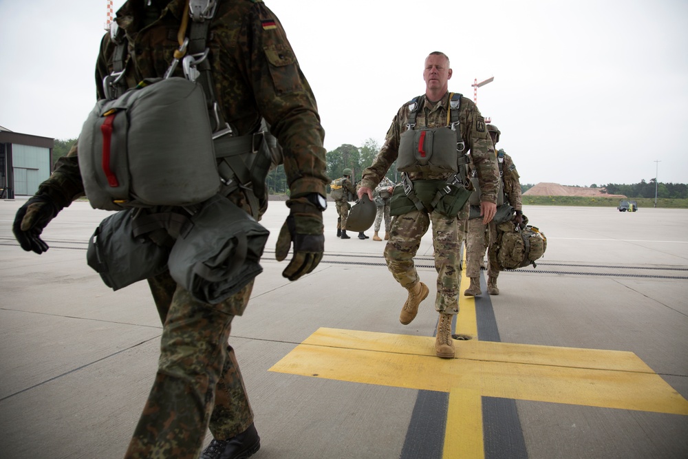 Military personnel in full parachute jump gear are walking across an airfield tarmac. The group includes at least one German soldier (identifiable by the German flag patch and Flecktarn camouflage uniform) and one or more U.S. Army soldiers in MultiCam/OCP uniforms. They appear to be preparing for or returning from a airborne/parachute operation at what appears to be a NATO or joint military airfield.