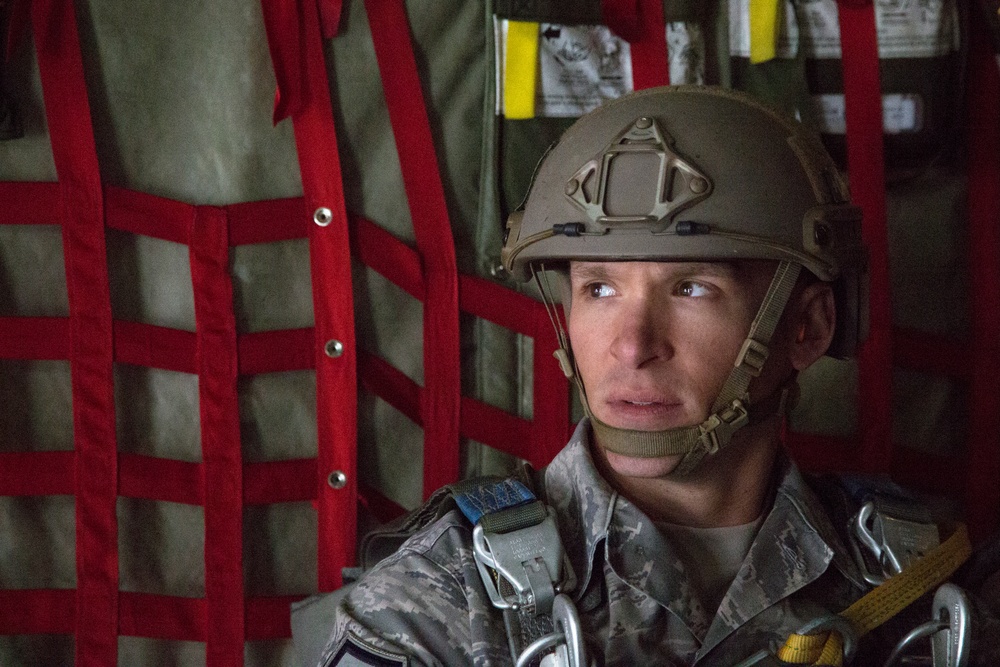 A military service member wearing a tan/coyote brown tactical helmet and digital camouflage uniform is seated inside what appears to be a military aircraft. The individual is equipped with parachute harness hardware including carabiners and straps, suggesting preparation for an airborne operation. The background shows the interior wall of the aircraft with red cargo netting and olive drab padding.