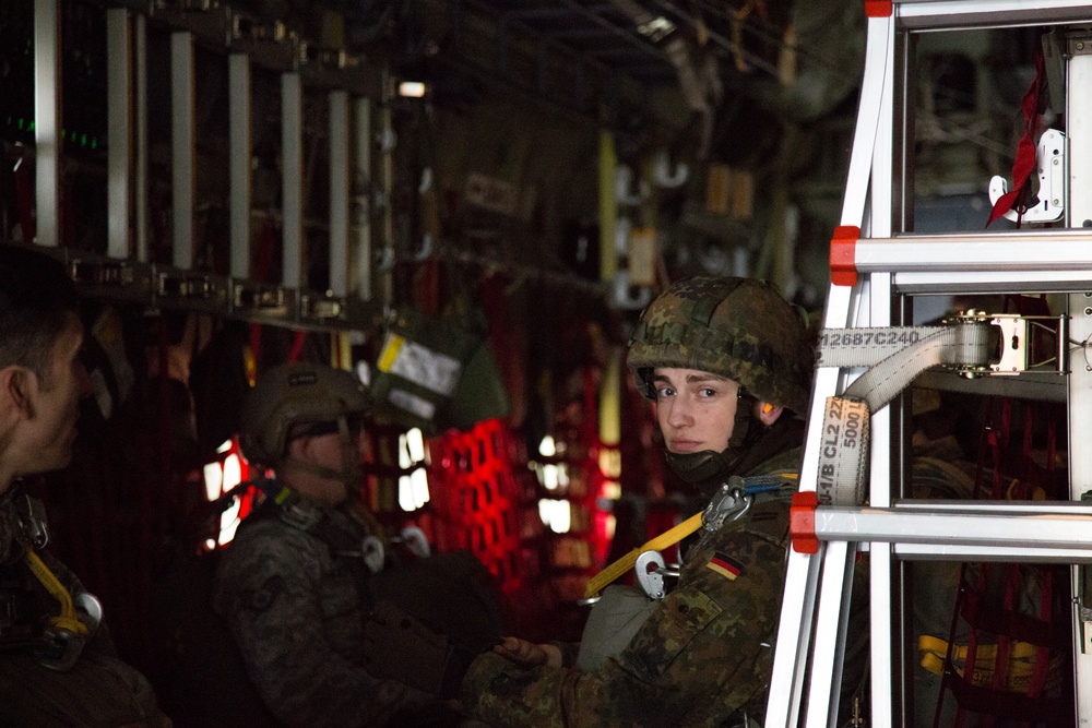 The image shows military personnel inside what appears to be a military transport aircraft cargo hold. A soldier wearing German Bundeswehr flecktarn camouflage uniform and helmet is prominently visible, holding or steadying a metal ladder or frame structure secured with ratchet straps. The interior is dimly lit with red ambient lighting typical of military aircraft night operations.