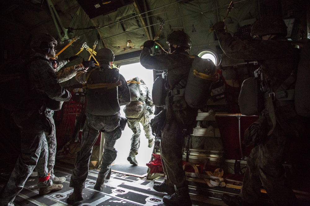 Military personnel are conducting a parachute jump operation from the open rear ramp of a military transport aircraft, likely a C-130 Hercules. At least one jumper is visible in the doorway preparing to or actively exiting the aircraft, while other personnel in full combat gear observe and assist. The interior lighting is dark with bright backlighting from the open ramp, creating strong silhouettes.