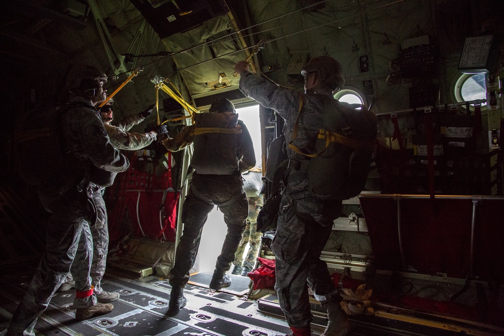 Military personnel are preparing to conduct a parachute jump from the open side door of a military cargo aircraft, likely a C-130 Hercules. Several soldiers in combat uniforms and helmets are visible near the jump door, with static lines attached overhead. The interior of the aircraft is dimly lit, contrasting sharply with the bright light visible through the open door.