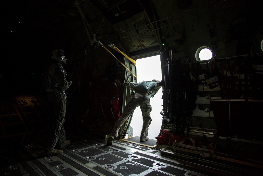 The image shows the interior of a military transport aircraft with two personnel near an open side door. One individual appears to be in the process of exiting or pushing equipment out of the aircraft door, while another stands and observes. The scene is dramatically lit with bright light flooding through the open door against the dark aircraft interior.
