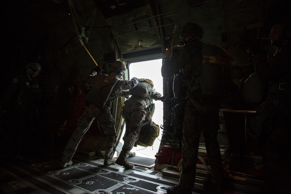Military personnel are conducting a parachute jump operation from the open rear door of a military transport aircraft, likely a C-130 Hercules or similar aircraft. One jumper is positioned at the door threshold in the process of exiting the aircraft, while other personnel assist and observe. The image is taken from inside the darkened cargo bay, with bright exterior light silhouetting the figures.