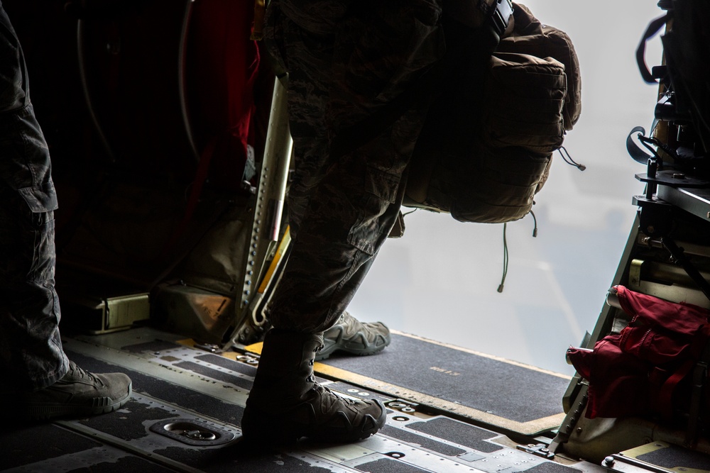 The image shows military personnel standing near the open rear ramp of a military transport aircraft, likely preparing for an airborne operation or aerial observation. One individual wearing camouflage uniform and carrying a large tactical backpack is leaning toward the open ramp edge. The scene is captured from inside the aircraft looking toward the open ramp with overcast sky visible outside.
