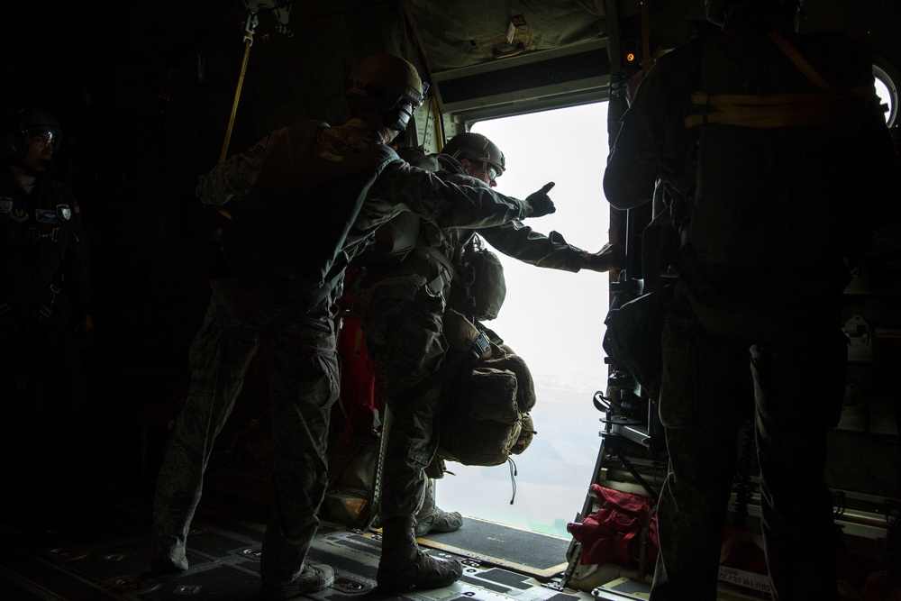 Military personnel are positioned at the open rear ramp of a cargo aircraft, appearing to prepare for a parachute jump operation. One individual is pointing outward through the open ramp while others stand equipped with helmets and large backpacks, likely parachute rigs. The scene is backlit by exterior light, creating strong silhouettes against the bright sky and water visible below.