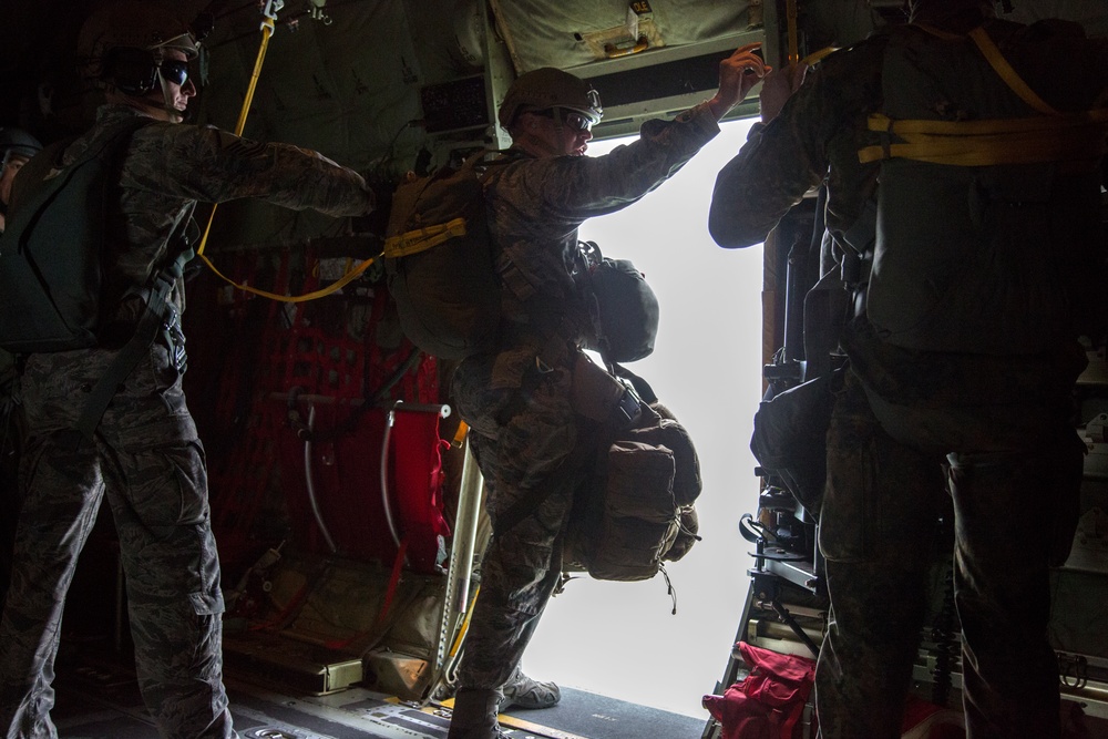 Military personnel in full combat gear and parachute equipment are positioned at the open door/ramp of a military transport aircraft, appearing to be preparing for a parachute jump. The interior of the aircraft is visible with yellow static lines, red equipment bags, and cargo straps. The bright exterior light contrasts sharply with the dark aircraft interior, creating a high-contrast silhouette effect.