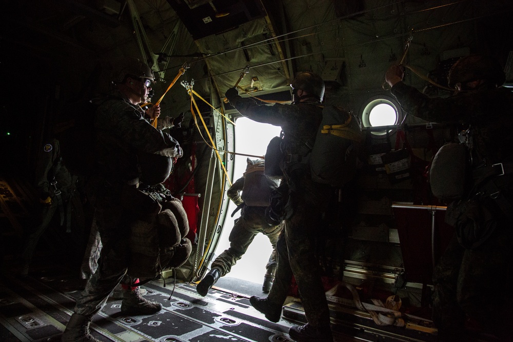 Military personnel are conducting a parachute jump operation from the open rear ramp of a military transport aircraft, likely a C-130 Hercules. One jumper is captured mid-exit through the open ramp door, while other personnel in full combat gear and parachute equipment stand by inside the aircraft. The interior is dark with strong backlighting from the open ramp, creating a high-contrast silhouette effect.