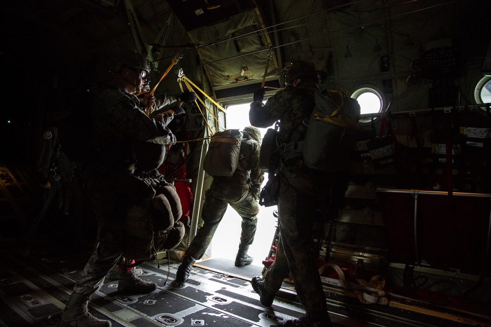 Military personnel are preparing to conduct a parachute jump from the open side door of a military transport aircraft, likely a C-130 Hercules. At least one jumper is positioned at the door threshold while others wait in line behind, with static lines attached to the overhead anchor cable. The interior is dimly lit with strong backlighting from the open door creating silhouetted figures.