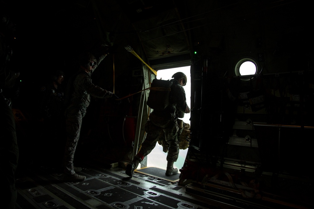 A military paratrooper is in the process of jumping from an aircraft through an open side door, silhouetted against bright exterior light. A jumpmaster or safety personnel stands to the left, holding a static line or safety strap. The interior of the military transport aircraft is visible, characterized by a dark cargo bay with a metal floor.