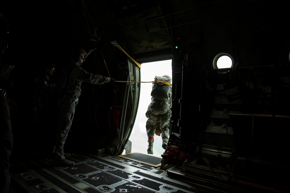 A military paratrooper is captured mid-exit jumping from the open door of a military aircraft, likely a C-130 or similar transport plane. A jumpmaster in camouflage uniform stands to the left, holding a yellow static line as the jumper exits. The image is taken from inside the dark aircraft cabin looking toward the bright exterior sky.