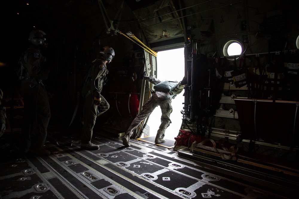 The image shows the interior of a military transport aircraft, likely a C-130 Hercules, with personnel conducting a parachute jump operation. One individual is actively exiting through the open side door while two others stand nearby in the cargo bay. The scene is dramatically lit by the bright exterior light contrasting with the dark aircraft interior.