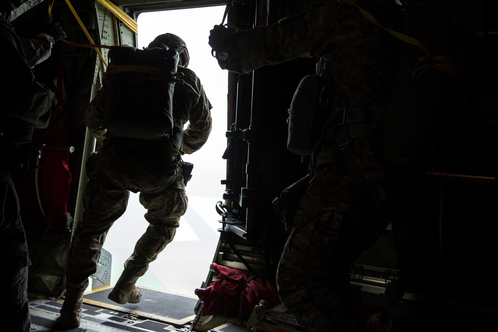 A military paratrooper is captured mid-jump exiting the open door/ramp of a military aircraft, likely a C-130 or similar transport plane. At least two other personnel in camouflage uniforms are visible inside the aircraft, one appearing to be a jumpmaster monitoring the exit. The image is taken from inside the aircraft looking toward the bright exterior, creating a strong silhouette effect.