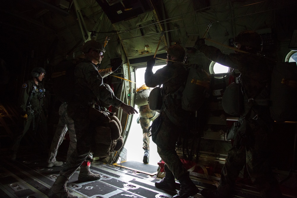 Military personnel are conducting a parachute jump operation from the open rear ramp of a military transport aircraft, likely a C-130 Hercules. At least one jumper is visible mid-exit at the ramp opening, while other personnel in full combat gear and parachute equipment stand inside the aircraft. The interior is dimly lit with bright light flooding in from the open ramp door.