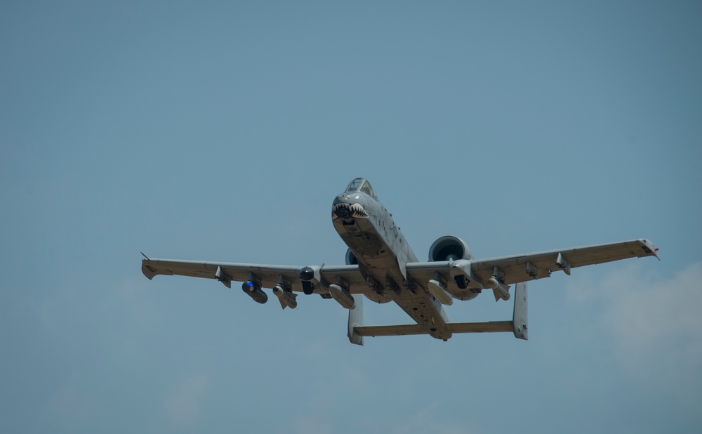 A U.S. Air Force A-10 Thunderbolt II (Warthog) attack aircraft is captured in flight against a hazy blue sky. The aircraft features a distinctive shark mouth nose art painted on the fuselage and appears to be carrying multiple underwing stores. The image is taken from a low angle, providing a frontal-slightly-below perspective of the aircraft.