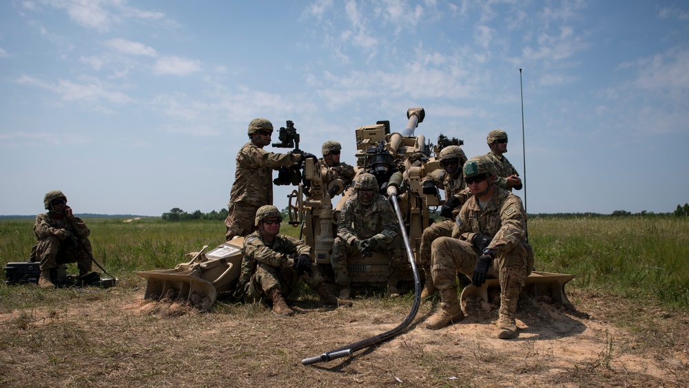 A group of military personnel in camouflage uniforms are gathered around what appears to be an M777 lightweight howitzer artillery piece in an open field. The soldiers appear to be conducting training operations, with some adjusting equipment on the weapon system while others rest nearby. The setting appears to be a military training range during daytime with clear weather conditions.
