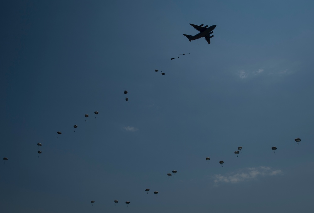 A large military transport aircraft is conducting a mass airborne drop operation against a blue sky backdrop. Numerous parachutes at various stages of deployment are visible descending below the aircraft, forming a trail pattern. The silhouetted aircraft and parachutes suggest a large-scale military airborne exercise or operation.