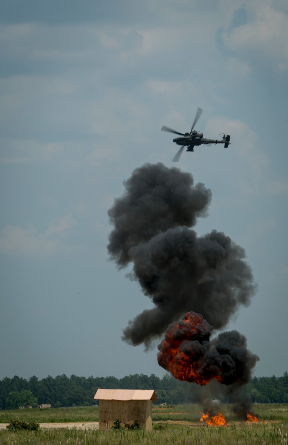 A military attack helicopter, consistent with an AH-64 Apache, is flying above a large explosion and rising black smoke column on an open field. The explosion produces orange-red flames and dense dark smoke, suggesting a strike on a ground target. A small wooden structure is visible in the foreground, likely a simulated target used during a training exercise or demonstration.