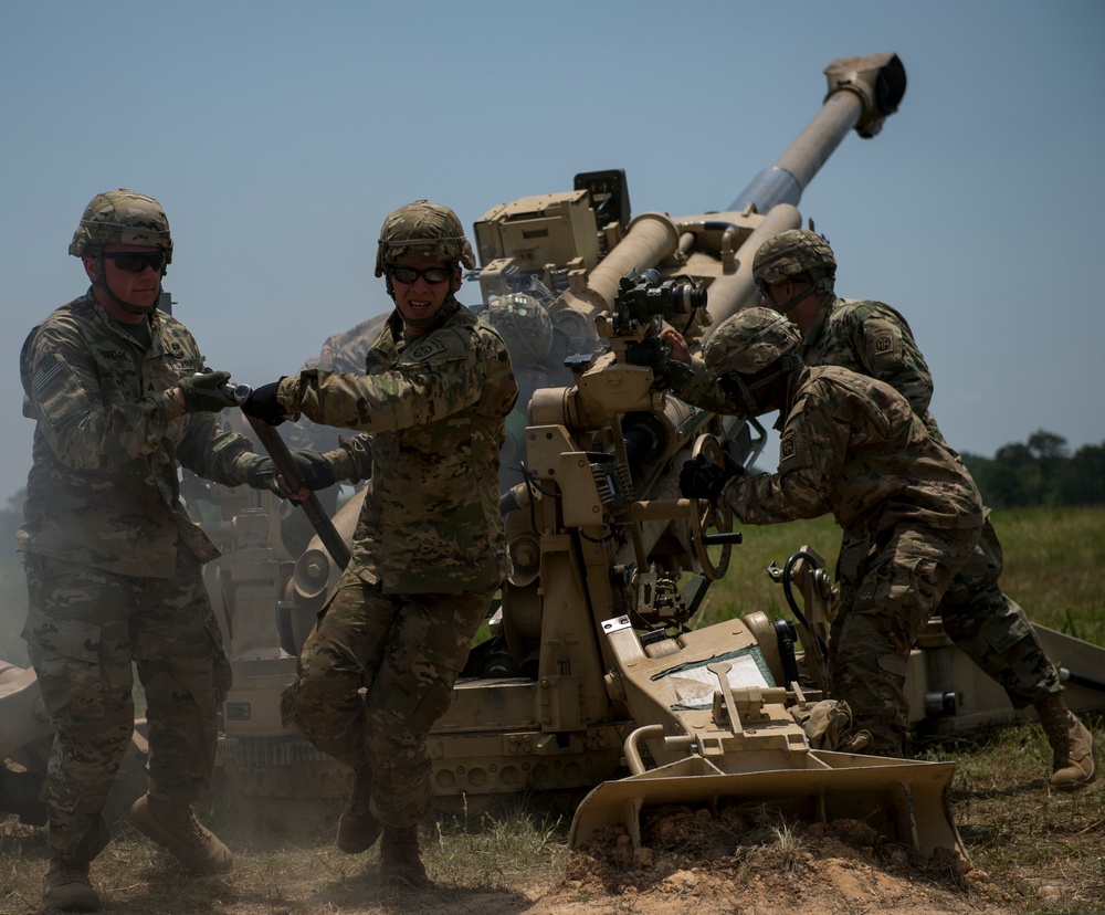 Three U.S. Army soldiers in MultiCam/OCP camouflage uniforms are operating an M777 155mm howitzer artillery piece in an open field. The soldiers appear to be in the process of loading or operating the howitzer, with visible smoke suggesting a recent or ongoing firing sequence. The setting appears to be a training exercise based on the controlled environment and daylight conditions.
