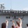 A group of children are waving up at a large naval warship, likely during a departure or homecoming event. At least one person visible on the ship's upper deck is waving back at the children below. The scene suggests a military send-off or welcome-back ceremony at a naval port.