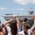 A group of young people, viewed from behind, are waving toward a large naval vessel — likely an aircraft carrier or amphibious assault ship — with personnel visible standing along its flight deck. The scene appears to depict a military homecoming or departure event at a naval port. Several individuals have their arms raised in greeting or farewell gestures directed at the ship's crew.