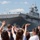 A large U.S. Navy amphibious assault ship is passing through a waterway, likely the Hudson River in New York City, during what appears to be Fleet Week. Civilians on shore are waving enthusiastically at the vessel, while crew members are visible standing on the flight deck. An American flag and what appears to be a Canadian flag are visible on the ship's superstructure, and a military helicopter is partially visible on the starboard side.