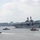 A large U.S. Navy amphibious assault ship is navigating a harbor waterway, escorted by multiple red-and-black tugboats. The scene appears to be in a major urban port, with a city skyline visible in the background. The ship features a flat flight deck characteristic of amphibious warfare vessels.